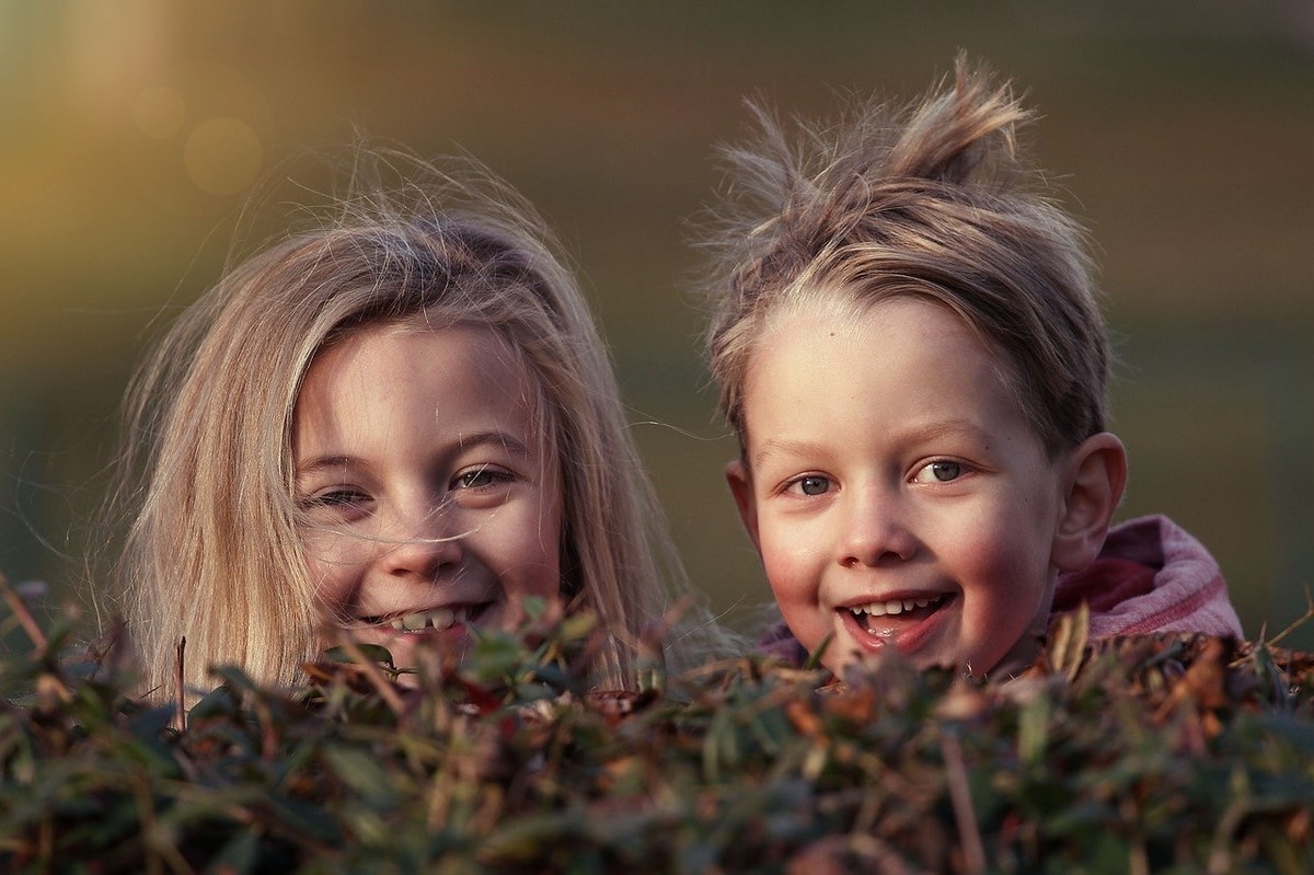 Zwei lachende Kinder schauen hinter einer Wiese hervor